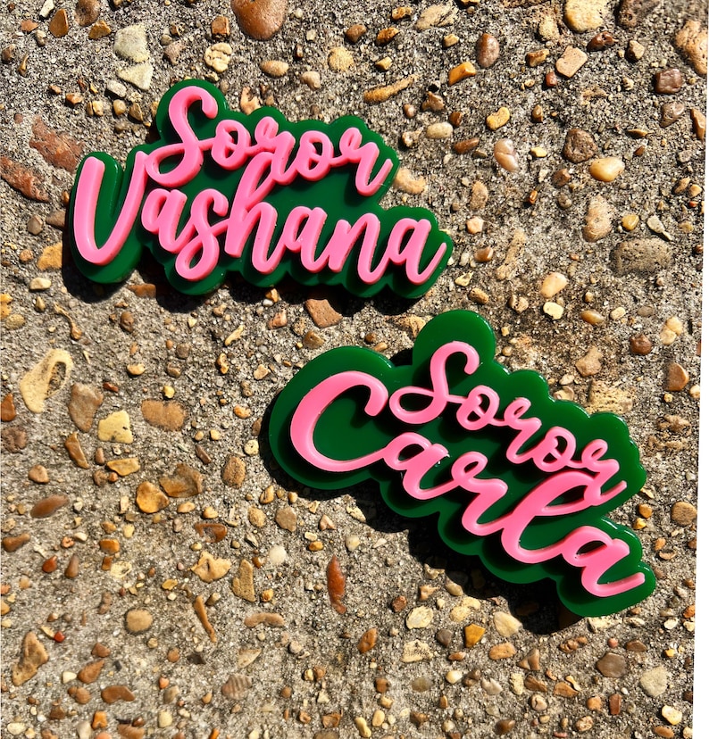 Op de afbeelding: Twee groene en roze acryl naamstickers met de tekst "Soror Vashana" en "Soror Carla".