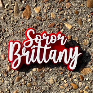 May include: Red and white acrylic sign with the text "Soror Brittany" in a cursive font.