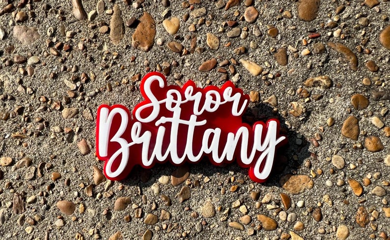 Op de afbeelding: Een rood en wit acryl bord met de tekst "Soror Brittany" in een cursief schrift.