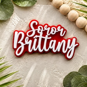 Peut inclure: Une plaque nominative personnalisée avec le texte Soror Brittany en police cursive blanche superposée sur un fond rouge uni. L'enseigne présente un aspect dimensionnel avec les lettres blanches en relief sur la base rouge contrastée.