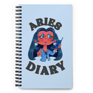 Puede incluir: Cuaderno de espiral azul claro con una ilustración de dibujos animados. La ilustración representa a una mujer con cabello azul sosteniendo un teléfono plegable. Las palabras "ARIES DIARY" están escritas en marrón.