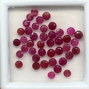 Könnte beinhalten: Eine Sammlung kleiner, runder, facettierter Rubine in verschiedenen Schattierungen von tiefem Rot und Pink. Diese losen Edelsteine sind in einer quadratischen weißen Aufbewahrungsbox mit einer weißen Schaumstoffeinlage angeordnet.