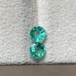 May include: Two round, faceted emerald gemstones with a vibrant green hue. The gems are displayed on a textured, light gray surface. The stones have a brilliant cut, reflecting light and showcasing their clarity.
