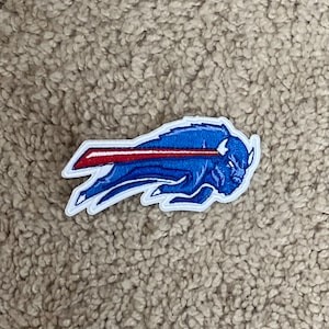 Puede incluir: Un parche bordado azul y rojo de un búfalo corriendo con una franja roja en la espalda. El parche está sobre una superficie marrón y peluda.