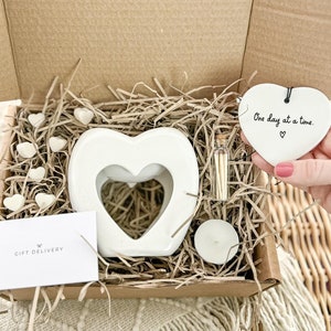 May include: A white ceramic heart-shaped wax melt burner with a smaller heart cut-out in the centre. The burner is surrounded by white wax melts and a small glass jar of matches. A white heart-shaped ceramic ornament with the text "One day at a time." is in the top right corner of the image.