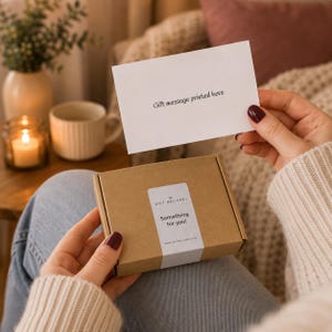Puede incluir: Una peque&ntilde;a caja de regalo rectangular de cart&oacute;n marr&oacute;n con una etiqueta blanca que dice Gift Delivery Something for you! y una tarjeta blanca encima con el texto Gift message printed here, sostenida por dos manos con esmalte de u&ntilde;as rojo oscuro.