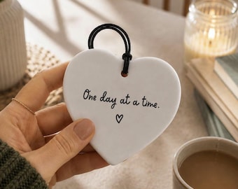 May include: A white heart-shaped ceramic ornament featuring the black printed text One day at a time. and a small heart symbol. The ornament has a smooth finish and includes a black cord loop at the top for hanging, providing a simple and clean design.