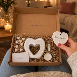 May include: A brown cardboard gift box containing a white iridescent heart-shaped wax warmer, a small glass vial of matches, a white tea light, eight small white heart-shaped wax melts, and a white heart-shaped ceramic tag with One day at a time.