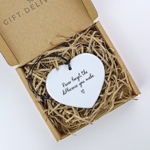 May include: White ceramic heart ornament with a black string for hanging. The heart has the text "Never forget the difference you make." written on it.