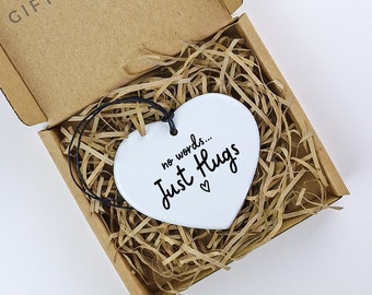 以下が含まれることがあります： 白いセラミック製のハート型のオーナメントで、黒い文字で「no words... Just Hugs」と小さなハートのシンボルが書かれています。オーナメントは、茶色の段ボール箱に詰められており、中には細かく切った紙が入っています。