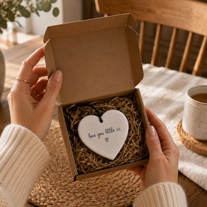 Puede incluir: Un adorno de cer&aacute;mica blanca en forma de coraz&oacute;n con un cordel negro, que presenta el texto impreso en negro "love you little sis." y un peque&ntilde;o icono de coraz&oacute;n. El adorno est&aacute; colocado dentro de una peque&ntilde;a caja de regalo de cart&oacute;n marr&oacute;n.