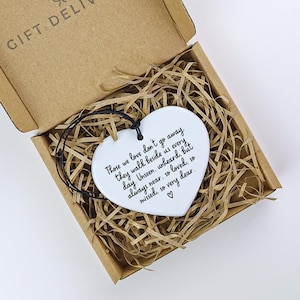 May include: A white heart-shaped ornament with a black string, inside a cardboard box filled with straw. The ornament has a handwritten quote: "Those we love don't go away..." A heartfelt gift.
