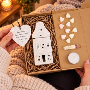 May include: A gift box containing a white ceramic house-shaped oil burner, ten small heart-shaped wax melts, a glass vial of matches, and a white tea light candle. A white heart-shaped ceramic ornament with the text New Home New Adventures New Memories Lily & George is held beside the box.