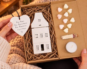 Puede incluir: Una caja de regalo que contiene un quemador de aceite de cerámica blanca con forma de casa, diez pequeñas ceras aromáticas en forma de corazón, un frasco de vidrio con cerillas y una vela de té blanca. Un adorno de cerámica blanca en forma de corazón con el texto New Home New Adventures New Memories Lily & George se sostiene junto a la caja.