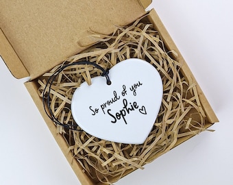 May include: White ceramic heart ornament with black text that reads "So proud of you Sophie" with a small heart symbol. The ornament is in a brown cardboard box with brown shredded paper.