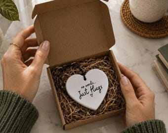 May include: A white heart-shaped ceramic ornament with a black string, featuring the printed text "no words... Just Hugs" with a small heart icon. The ornament is nestled inside a small, open brown cardboard gift box filled with shredded paper.