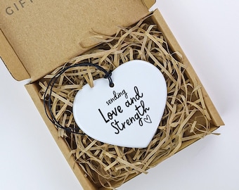 以下が含まれることがあります： 白いセラミック製のハート型のオーナメントで、黒い文字で「sending love and strength」と書いてあり、小さなハートのシンボルが付いています。オーナメントは、茶色の段ボール箱の中に、細かく切った紙と一緒にあります。