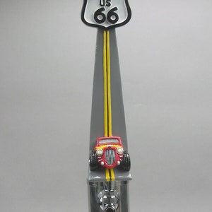May include: A silver beer tap with a Route 66 sign and a red hot rod car on top. The sign is white with black lettering and the car has yellow headlights.