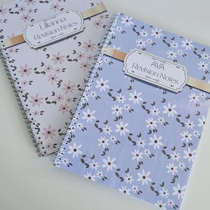 May include: Two floral-patterned spiral-bound notebooks. One has a light purple cover with pink flowers, the other a light blue cover with white flowers. Each notebook features a label that reads "Revision Notes".