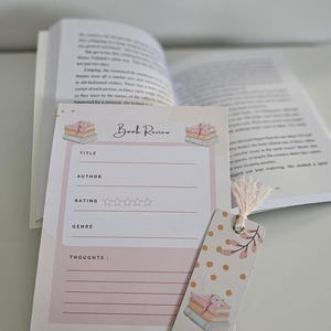 May include: A pink and white book review notepad and bookmark with a floral design. The notepad has spaces for title, author, rating, genre, and thoughts. An open book is visible in the background.