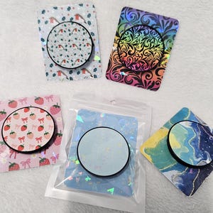 May include: A selection of phone grips with diverse designs. One has a strawberry pattern, another a rainbow swirl, and others with bird, marble, and abstract patterns. Each grip has a black circular base.