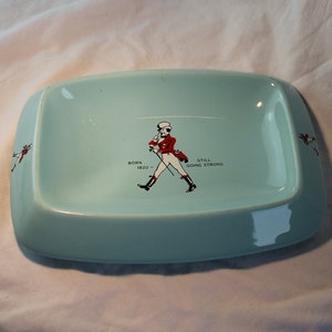 May include: A light blue ceramic ashtray with a white border. The centre features a black and white illustration of a man in a red coat and white trousers walking with a cane. The text "Born 1820 - Still Going Strong" is printed below the illustration.