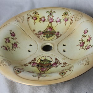 Puede incluir: Una pantalla de lámpara vintage de cerámica de color crema con un diseño floral. La pantalla tiene cuatro secciones con un agujero central para la fijación de la lámpara. El diseño floral presenta rosas rosas, hojas verdes y detalles dorados.