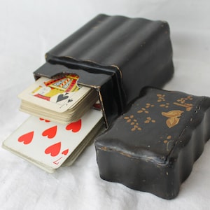 Puede incluir: Una caja de madera negra con una baraja de cartas dentro. La caja tiene un diseño decorativo en la parte superior y está ligeramente abierta. Las cartas son visibles e incluyen un Jack de picas y un 7 de corazones.