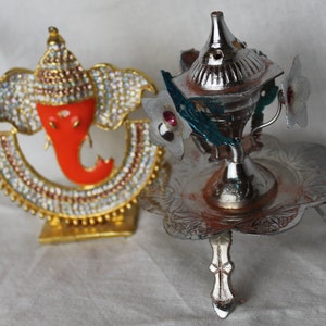 May include: Two decorative religious items. One is a gold-colored Ganesh statue with an orange face and jeweled accents. The other is a silver incense burner with floral embellishments and a decorative base.