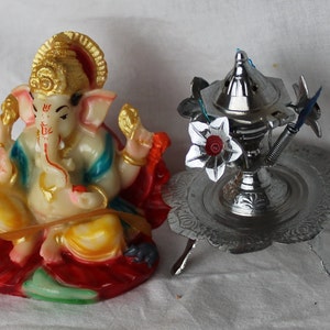 May include: A colorful, ornate statue of the Hindu deity Ganesha, sitting on a red and green lotus base.  A silver incense burner with a floral design sits on a silver stand with three legs.