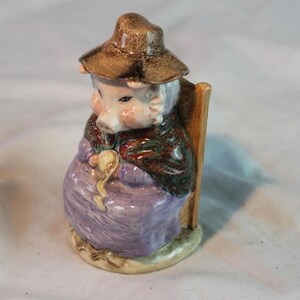 May include: A ceramic figurine of a pig-like character wearing a brown hat, a patterned shawl, and a purple dress. The figurine is seated in a wooden chair and holds a gold-coloured object. The base is a light tan colour.