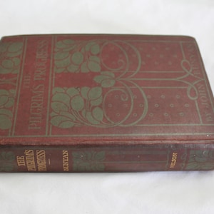May include: A vintage copy of "The Pilgrim's Progress" by John Bunyan. The book has a burgundy cover with green decorative elements and text. The book is closed and lies on a white surface.