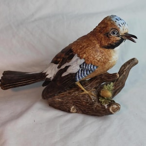 May include: A detailed ceramic bird figurine, featuring a brown body, white and blue wing accents, and a blue-grey head. The bird is perched on a brown, textured branch with a small acorn. The figurine is a decorative item.