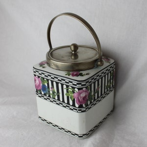 May include: A white ceramic biscuit jar with a black and white striped design and floral accents. The jar has a silver lid with a brass handle.