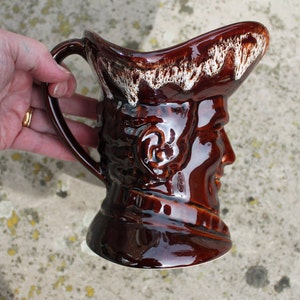 Könnte beinhalten: Ein brauner Keramik Krug mit einem weißen Glasurdesign. Der Krug ist in Form eines menschlichen Kopfes mit Hut. Der Krug hat einen Griff und einen Ausguss.