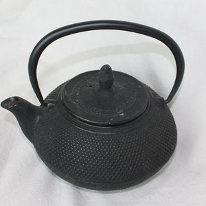 Könnte beinhalten: Eine schwarze gusseiserne Teekanne mit einer strukturierten Oberfläche und einem Deckel mit einem kleinen Knopf oben. Die Teekanne hat einen gebogenen Griff.