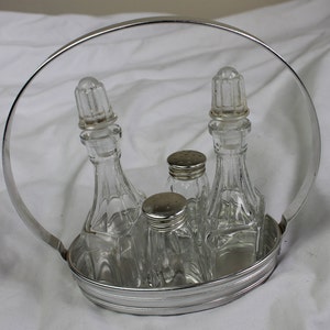 May include: A silver metal condiment holder with a circular base and a handle. The holder contains four glass bottles with stoppers. The bottles are clear and have a textured surface.