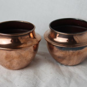 Puede incluir: Dos ollas de cobre con forma redondeada y un borde ligeramente acampanado. Las ollas tienen un acabado brillante y pulido.