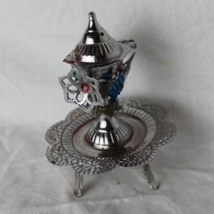 May include: Silver incense burner with a decorative flower design and blue accents. The burner sits on a three-legged stand with a scalloped edge.