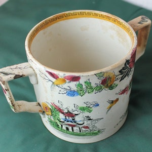 Op de afbeelding: Een witte keramische mok met twee handvatten, met een kleurrijk bloemen- en vogelontwerp met een gele rand rond de bovenkant.