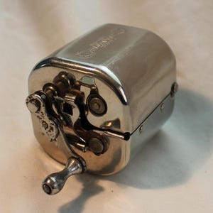 May include: A silver metal pencil sharpener with a crank handle. The sharpener has a small metal chain and gears visible on the side.