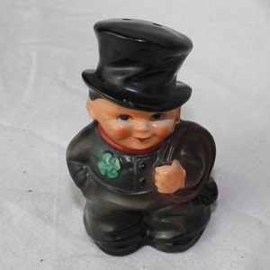 May include: A ceramic figurine of a chimney sweep wearing a black top hat and holding a black bag. The figurine has a smiling face and a green shamrock on its chest.