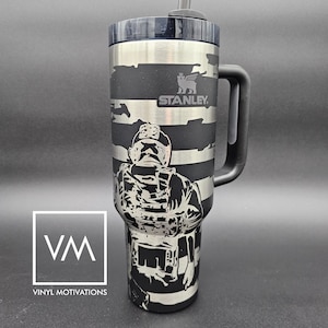 May include: A black and silver stainless steel travel mug with a black handle. The mug has a camouflage pattern and a graphic of a soldier with a rifle. The mug has the text "STANLEY" on it.