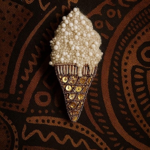Può includere: Una spilla artigianale a forma di cono gelato con una parte superiore testurizzata composta da piccole perline bianche e una base a cono decorata con paillettes dorate metallizzate e perline tubolari color bronzo.