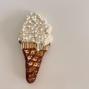 May include: A beaded ice cream cone brooch featuring a scoop of white seed beads and a cone crafted from brown thread, gold-toned sequins, and metallic tube beads. The design includes a small drip of white beads cascading down the side.
