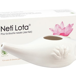 May include: A white ceramic Neti Lota nasal rinse pot with a spout and handle, in front of a box with the text "Neti Lota" and a pink lotus flower illustration. The box also has French text.