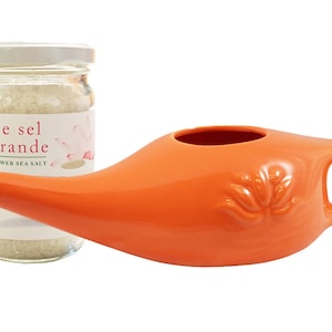 Op de afbeelding: Een oranje keramische neti pot met een handvat en een tuit, naast een glazen pot Fleur de sel de Guérande zeezout. De pot heeft een witte deksel en een etiket met roze bloemaccenten. De neti pot heeft een decoratief bloemmotief.