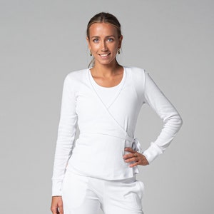 Op de afbeelding: Een witte wikkel top met lange mouwen en een striksluiting, gecombineerd met een witte broek. De top heeft een gelaagd ontwerp en is gemaakt van een zacht, comfortabel materiaal. De outfit is geschikt voor casual of professionele kleding.