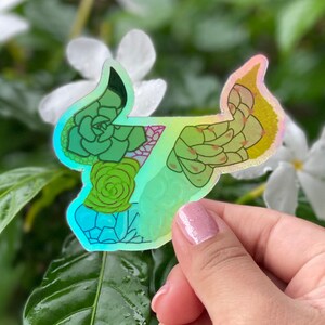 May include: A holographic sticker featuring a green succulent design in the shape of the number seven. The sticker has a rainbow iridescent finish.