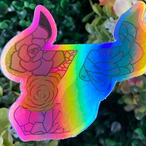May include: A holographic sticker with a rainbow background featuring a floral design of succulents. The sticker is outlined in black and has a glossy finish.